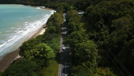 Modern-silver-car-driving-on-a-road-along-the-seaside-coast-with-sandy-beach-and-lush-green-tree-forest-and-blue-water-sea