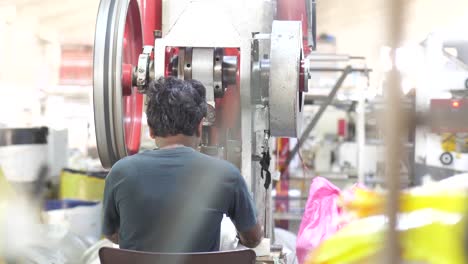 View-Behind-Male-Pakistani-Worker-Sorting-Plastic-Bags-At-Desk-With-Large-Spinning-Belt-Pully-Wheel-In-Factory-In-Karachi