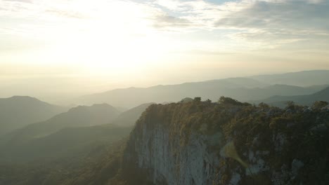 Aerial-view-of-sunrise-in-the-mountains,-soar-above-the-steep-cliffs-towards-the-suns-rays