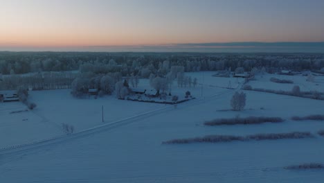 Aerial-establishing-shot-of-a-rural-landscape,-countryside-house,-agricultural-fields-and-trees-covered-with-snow,-cold-freezing-weather,-golden-hour-light-glowing,-wide-drone-shot-moving-forward