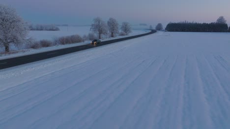 Aerial-establishing-shot-of-a-rural-landscape-with-road-and-car,-agricultural-fields-and-trees-covered-with-snow,-cold-freezing-weather,-golden-hour-light,-wide-ascending-drone-shot-moving-forward