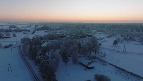 Aerial-establishing-shot-of-a-rural-landscape,-countryside-house,-agricultural-fields-and-trees-covered-with-snow,-cold-freezing-weather,-golden-hour-light-glow,-wide-drone-shot-moving-forward-high