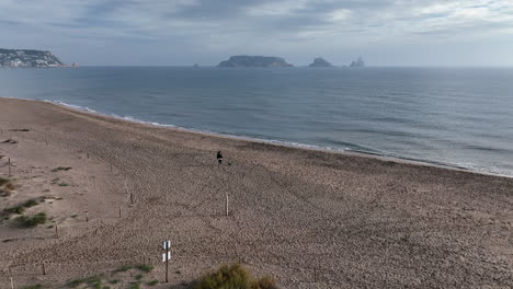 Aerial-of-woman-and-her-dog-running-alone-in-beach,-Medes-Islands-in-background