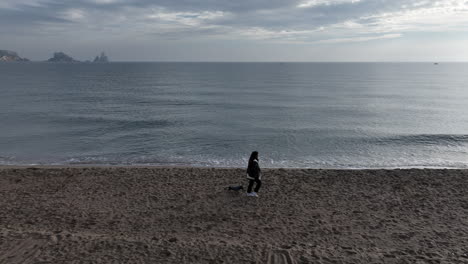 Tracking-aerial-shot-of-woman-walking-small-dog-on-empty-beach,-cloudy-day