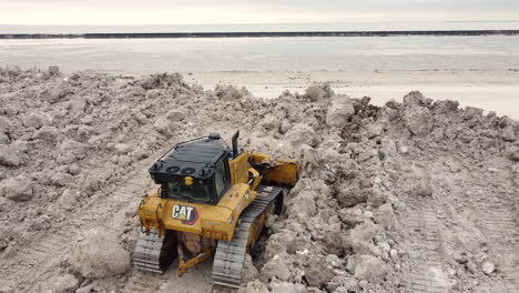 Close-aerial-of-bulldozer-moving-snow-by-lake-after-blizzard,-Buffalo