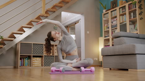 Young-fit-woman-sits-cross-legged-on-yoga-mat-at-home-stretching-with-arms-out