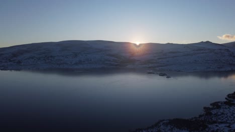 Long-Lagoon-Dam-Flyover,-Sunset-Flare-Picking-Through-Snow-Covered-Hill,-Serra-Da-Estrela