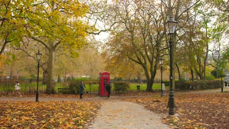 People-Walking-By-Red-Telephone-Booth-At-The-Park-In-London,-UK-With-Autumn-Colors