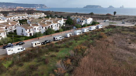 Orbiting-aerial-view-row-of-parked-campervans-on-Estartit-waterfront-overlooking-Medes-islands-seascape