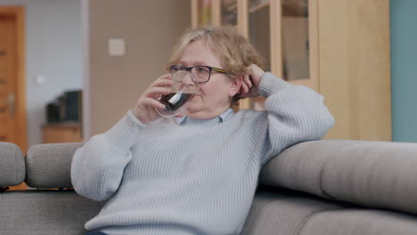 Elderly-caucasian-woman-with-glasses-relaxes-on-sofa-with-a-beverage