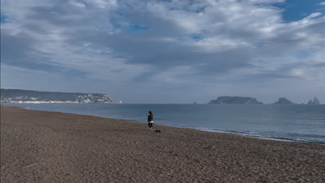 Woman-walking-miniature-Dachshund-across-empty-beach-overlooking-Medes-islands-coastline-at-sunrise