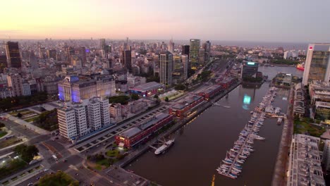 Aerial-view-dolly-in-of-the-docks-that-currently-have-luxury-restaurants-and-the-entrance-with-the-river-dike,-blue-hour-with-the-city-and-its-illuminated-buildings-in-Puerto-Madero