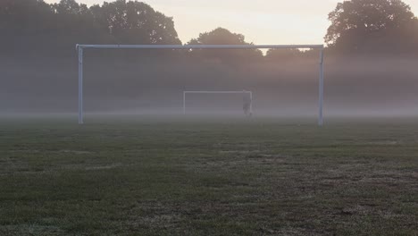 Man-jogging-past-football-goal-posts-in-heavy-fog,-with-his-pet-dog