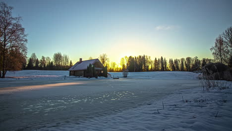 Timelapse-of-majestic-sunrise-in-the-background-of-a-traditional-small-wooden-house-in-the-snowy-winter-landscape-and-clouds-overhead,-sunlight-falling-on-the-ground