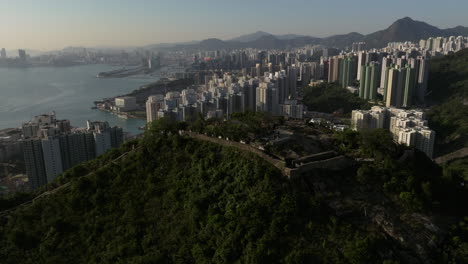 Orbiting-shot-of-Devil's-peak-with-Victoria-Harbour,-Kowloon-Bay-and-Mountain-range-in-backdrop-during-golden-hour,-Hong-Kong