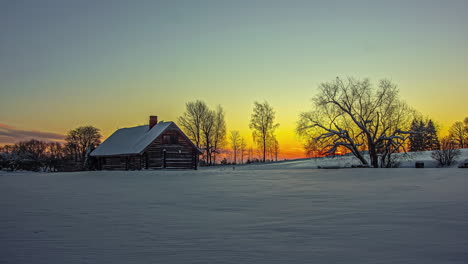 Static-shot-of-beautiful-wooden-cottage-covered-with-thick-layer-of-white-snow-with-sun-rising-in-timelapse-in-the-background-during-sunrise