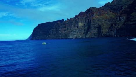 Small-boat-cruising-Open-Blue-ocean-In-Front-of-high-Rise-Rocky-Mounntains,-Spain