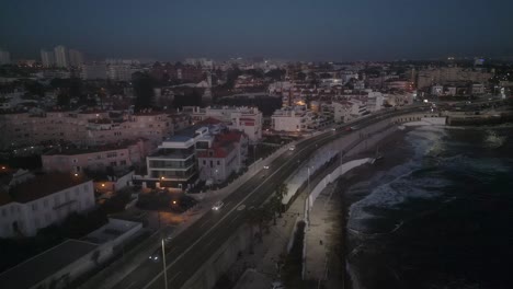 Aerial-view-of-a-small-town-almost-at-night-in-Cascais-with-some-traffic-and-small-waves-crashing-on-shore,-near-Lisbon,-on-the-coast-of-Portugal