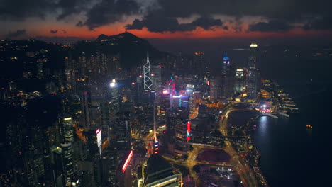 Ultra-Wide-slow-tracking-backwards-aerial-drone-shot-of-suspenseful-harbour-front-skyline,-Hong-Kong,-with-moody-cinematic-sunset-afterglow