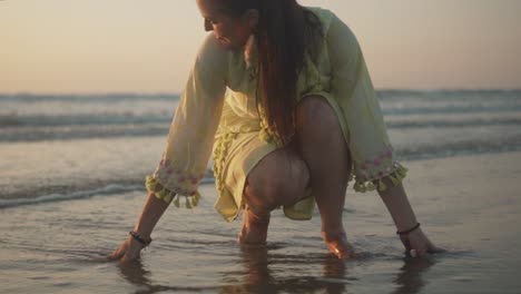 Woman-in-a-light-yellow-dress-and-brown-bracelets-on-both-hands-squatting-and-looking-at-the-shore-with-her-hands-in-the-water