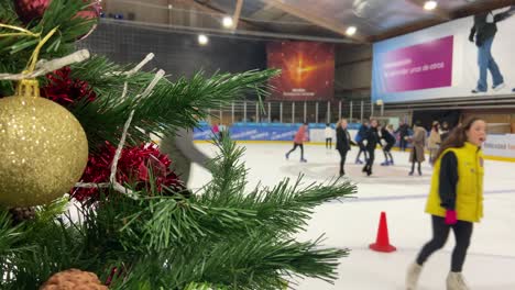 Close-up-of-Christmas-tree-with-many-people-ice-skating-in-indoor-rink-in-background