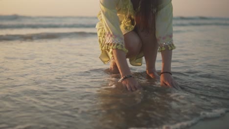Frau,-Die-Am-Ufer-Hockt-Und-Mit-Wasser-Spielt,-Während-Kleine-Schäumende-Wellen-über-Den-Sand-Spülen