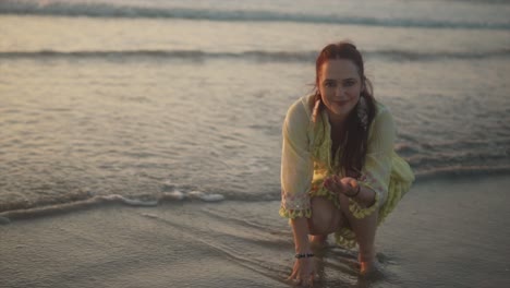 Woman-looking-at-the-camera-smiling-while-squatting-at-the-shoreline-with-one-hand-in-the-sand-and-another-holding-a-small-object