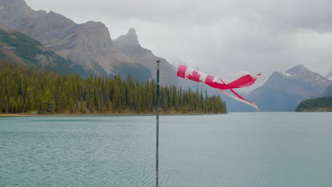 Kanadische-Flagge,-Die-Mit-Dem-Wind-In-Der-Nähe-Eines-Atemberaubenden-Sees-Mit-Grünem-Wasser-Aufwacht,-Der-Von-Hohen-Bergen-In-Den-Kanadischen-Rocky-Mountains-Umgeben-Ist