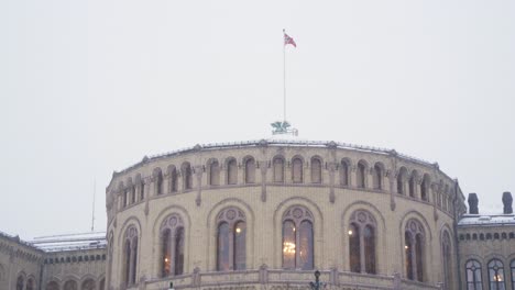 Norwegische-Nationalflagge,-Die-An-Kalten,-Verschneiten-Tagen-über-Dem-Parlamentsgebäude-In-Oslo-Weht,-An-Dem-Menschen-Vorbeigehen
