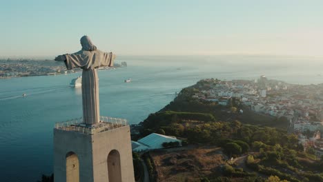 Drone-footage-captures-Jesus-Christ-statue-in-Lisbon,-Portugal,-showcasing-its-size-and-location-on-a-hill-surrounded-by-the-city-and-greenery