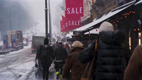 View-Looking-Along-Karl-Johan-Shopping-Street-With-People-Walking-Past-Restaurants-And-Sale-Signs-During-Heavy-Snow-Fall-In-Oslo