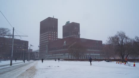 Oslo-City-Hall-On-Cloudy-Overcast-Winter-Day-With-Snow-Falling-With-People-Walking-Past-Viewed-From-Radhusplassen