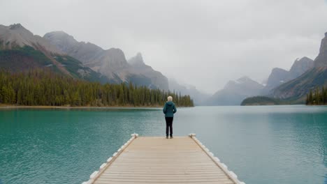 Outdoorsy-woman-looking-mountain-the-horizon-on-the-edge-of-a-small-pier-floating-on-a-stunning-green-water-glacial-lake-nestled-in-the-Canadian-rocky-mountains