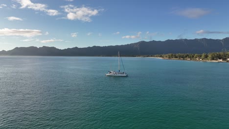 60-fps-drone-aerial-footage-of-hawaii-ocean-with-catamaran-in-blue-water-with-Waimanalo-mountains-in-the-background-and-lots-of-trees-midday-blue-skies