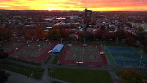 Aerial-tilt-up-reveal-from-basketball-courts-in-downtown-city-park-to-skyline-of-small-American-city-during-beautiful-autumn-sunset-in-USA