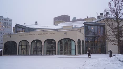 Schnee-Fällt-Vor-Dem-Nobelfriedenszentrum-In-Oslo,-Norwegen,-Mit-Zwei-Personen-Draußen-Und-Dann-Vorbeigehen
