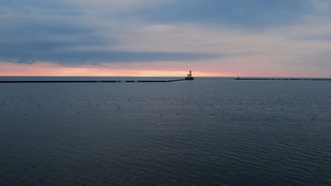 aerial-flying-low-above-water-Lake-Michigan-towards-Milwaukee-breakwater-light,-captured-at-sunrise,-historical-landmark-in-Wisconsin,-cinematic-shot-with-birds-on-lake