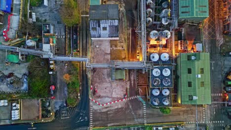 Forward-moving-aerial-footage-of-a-large-industrial-plant-at-dusk,-showing-pipework-structures,-buildings,-cooling-towers,-steam,-and-work-vehicles