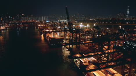 Dunkle-Portalkräne-Mit-Leuchtenden-Flutlichtern-Und-Containerschiff-In-Betrieb-An-Einem-Terminal-Mit-Der-Skyline-Von-Hongkong-Im-Hintergrund
