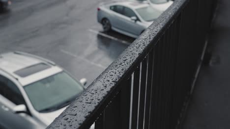 A-close-up-shot-of-rain-dripping-onto-a-metal-handrail-for-a-walkway-with-a-parking-lot-in-the-background