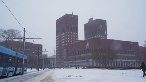 Oslo-City-Hall-On-Cloudy-Overcast-Winter-Day-With-Snow-Falling-With-Tram-Going-Past-Viewed-From-Radhusplassen