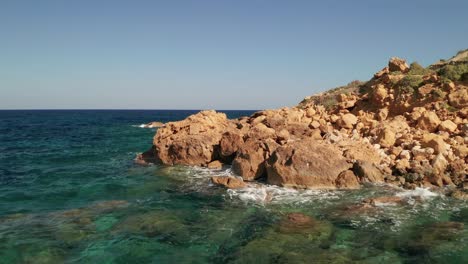 Aerial-forward-moving-shot-of-a-seashore-with-beautiful-turquoise-water-of-the-Mediterranean-Sea-splashing-on-the-red-coloured-rocks