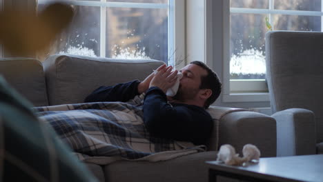 Man-with-flu-sneezing-into-napkin-while-lying-down-on-sofa-in-living-room