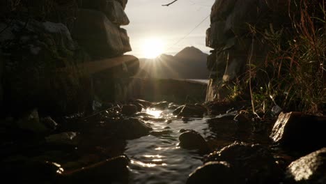 The-camera-slowly-tilts-down-to-reveal-the-sun-setting-behind-a-mountain-in-the-background-as-a-small-river-gently-flows-under-an-arch-in-a-stone-wall