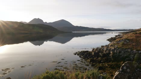 The-camera-gently-tilts-to-reveal-the-sun-reflecting-off-of-the-surface-of-a-tidal-sea-loch-in-the-north-west-Highlands-of-Scotland-just-before-setting-behind-mountains