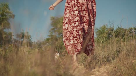 A-slow-motion-shot-of-an-attractive-Caucasian-female-wearing-a-red-floral-dress,-happily-skipping-and-running-along-a-path-carefree-in-a-field-outdoors,-India