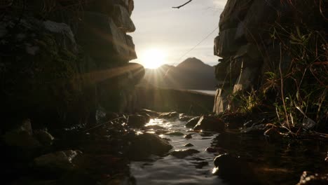 The-the-sun-sets-behind-a-Scottish-mountain-to-produce-a-sunburst-in-the-background-as-a-small-river-gently-flows-under-an-arch-in-a-stone-wall