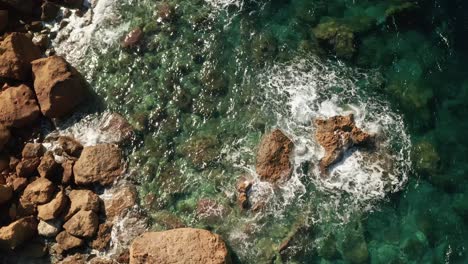 Aerial-tilt-down-reveal-shot-of-a-shore-with-beautiful-red-rocks-and-clear-turquoise-water-and-waves-crushing-on-rocks-on-the-beach-near-Rampla-Beach-on-the-island-of-Gozo,-Malta