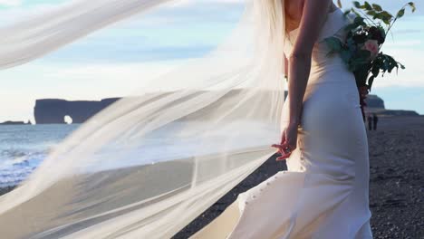 Long-Wedding-Bridal-Veil-Flowing-In-The-Wind-Behind-Caucasian-Bride-In-White-Dress-Standing-On-Black-Sand-Beach-During-Sunset