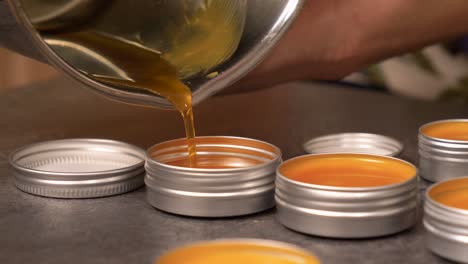 Close-up-shot-of-pouring-a-hot-yellow-orange-liquid-beeswax-balm-into-an-empty-circular-aluminum-container